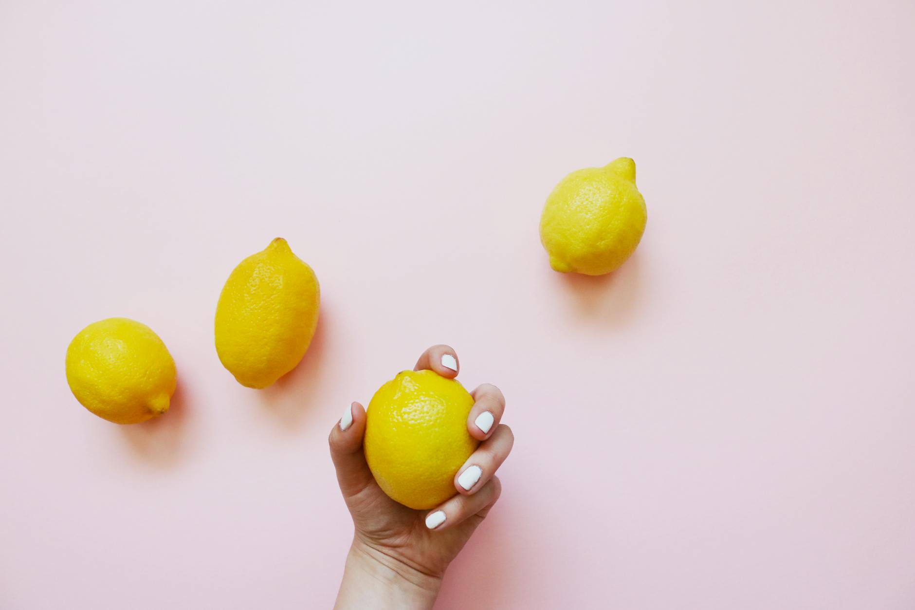 Una mano con unghie bianche tiene un limone su un delicato sfondo rosa, circondata da altri limoni