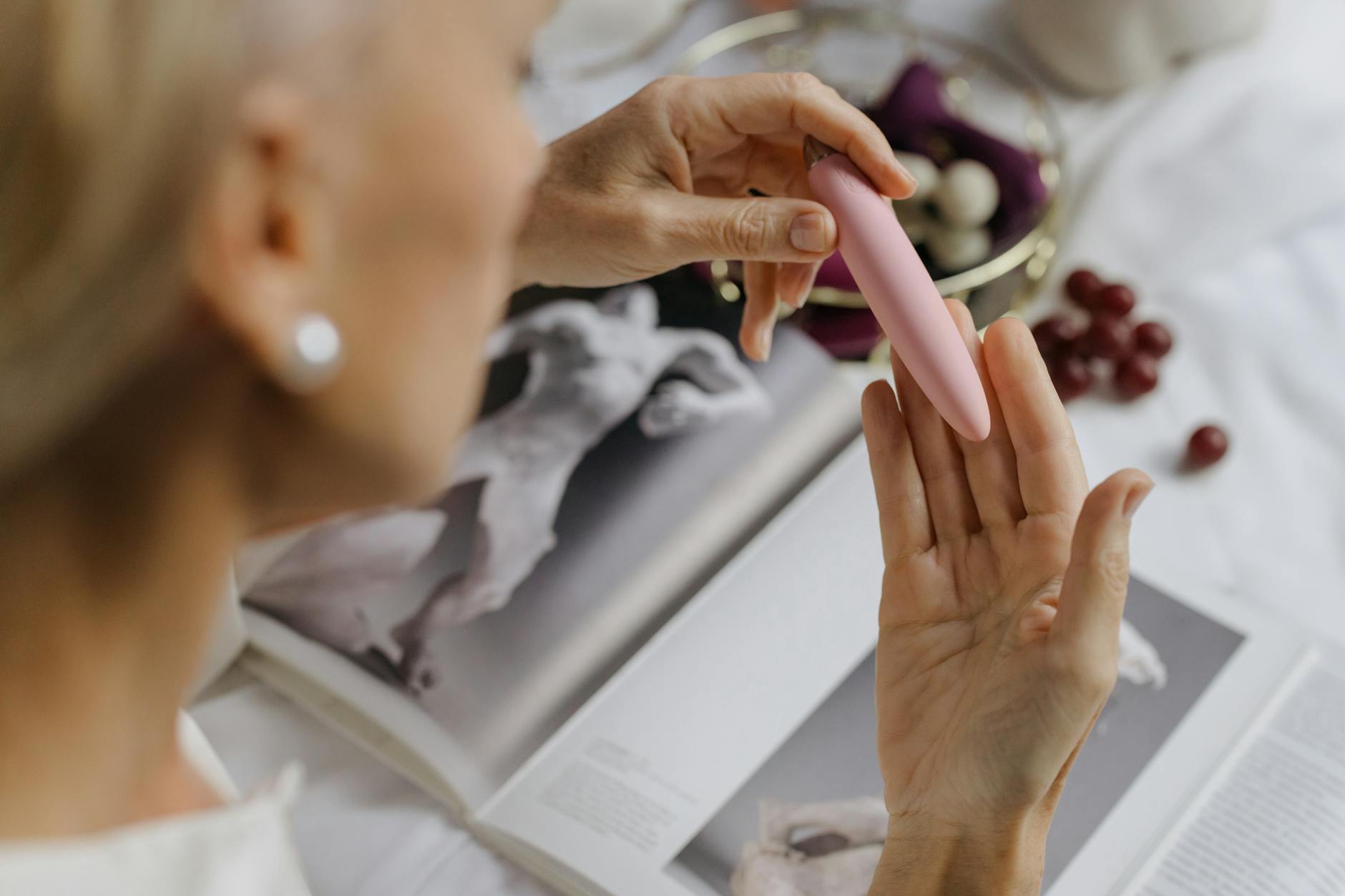 Primo piano di una donna che tiene un vibratore rosa con libri d'arte sullo sfondo, enfatizzando temi intimi e personali.